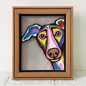 May include: A framed colorful paper art piece of a dog's face peeking out from behind a chevron pattern. The dog's face is made of layered paper in shades of pink, blue, green, yellow, and brown.