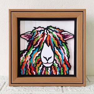 May include: A framed paper art piece of a sheep with a colorful, layered, wavy fleece. The sheep's face is white with black eyes and a pink nose.