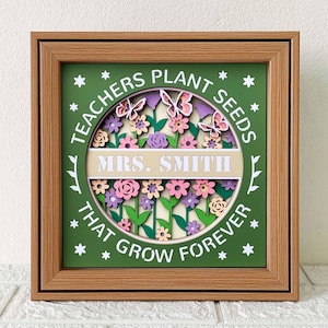 May include: A green framed wooden wall art with a layered paper cut design of flowers, butterflies, and leaves. The text reads "Teachers Plant Seeds That Grow Forever" and "Mrs. Smith".