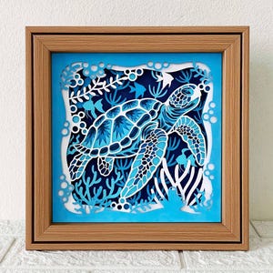 May include: A framed paper art piece depicting a blue and white sea turtle swimming in a blue and white coral reef. The turtle is cut out of paper and layered to create a 3D effect.
