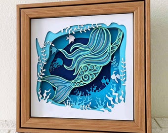 May include: A papercraft mermaid with blue and green tones, swimming through a blue and white ocean scene. The mermaid is surrounded by seaweed and jellyfish. The scene is framed in a wooden frame.