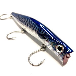 Puede incluir: Un señuelo de pesca con cuerpo azul oscuro y plateado que presenta un patrón de rayas oscuras similar al de una caballa. El señuelo tiene un acabado reflectante, un ojo prominente con pupila negra y tres anzuelos triples de metal.