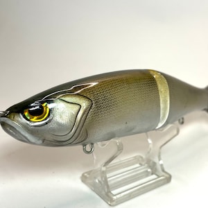 May include: A silver and brown fishing lure with a white stripe and a yellow eye. The lure is on a clear plastic stand.