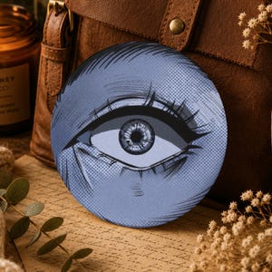 Può includere: Un sottobicchiere circolare con un'illustrazione in stile manga dai toni blu di un singolo occhio con ciglia dettagliate e ombreggiatura a punti. Il design grafico cattura un'iride e una pupilla stilizzate, presentate con un'estetica da fumetto.