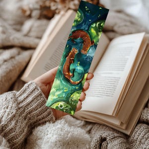 May include: A rectangular bookmark featuring a digital illustration of two brown otters swimming in deep blue water surrounded by vibrant green lily pads and aquatic foliage, with a small white circular logo at the bottom edge of the design.