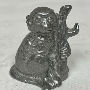 May include: A small, silver-colored metal figurine of a monkey sitting upright. The monkey is depicted with a textured surface, holding a vertical branch or tree trunk structure on its right side. The figure has a rounded head and defined facial features.