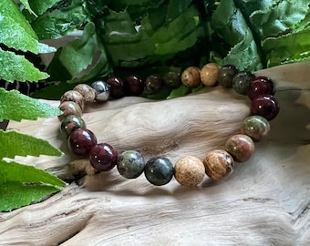 May include: A bracelet made of natural stone beads in shades of red, green, brown, and tan. The beads are strung on an elastic cord. The bracelet is displayed on driftwood, with green foliage in the background. The beads are approximately 1 cm in diameter.
