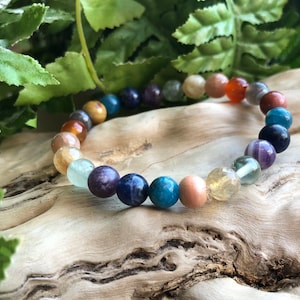 May include: A colourful beaded bracelet with stones in shades of green, blue, brown, and orange. The bracelet is made of natural stones and is resting on a piece of driftwood.