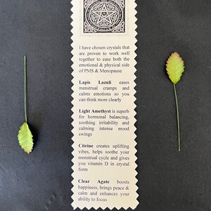 May include: A cream-colored card with a pentagram design and text about crystals for PMS and menopause, surrounded by four green leaves with red tips. The card lists crystals like Lapis Lazuli and Citrine.