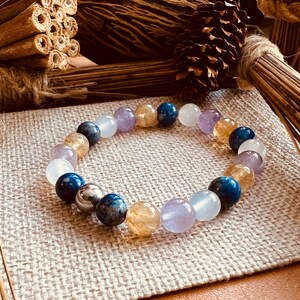 May include: A beaded bracelet featuring a variety of colorful gemstones, including blue, yellow, purple, and white beads. The bracelet is displayed on a neutral linen surface, with natural decorative elements in the background.