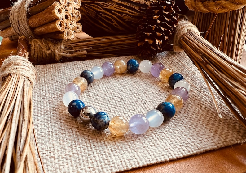 May include: A beaded bracelet featuring a variety of colorful gemstones, including blue, yellow, purple, and white beads. The bracelet is displayed on a neutral linen surface, with natural decorative elements in the background.