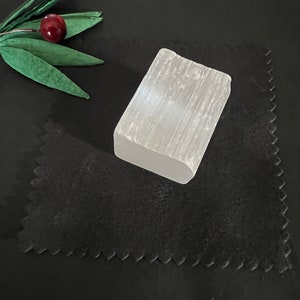 May include: A rectangular, translucent white selenite crystal sits on a black surface. The crystal has a rough, natural texture. A decorative sprig with green leaves and a red berry is in the upper left corner.