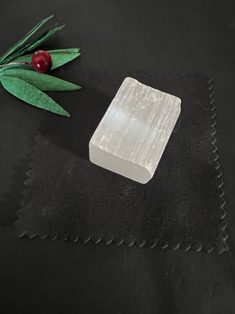 May include: A rectangular, translucent white selenite crystal sits on a black surface. The crystal has a rough, natural texture. A decorative sprig with green leaves and a red berry is in the upper left corner.