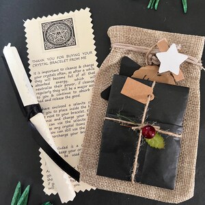 May include: A gift set with a burlap bag, wrapped package, and a scroll. The wrapped package is black with twine and a red berry. The scroll is tied with a black ribbon. The text on the card reads "THANK YOU FOR BUYING YOUR CRYSTAL BRACELET FROM ME."
