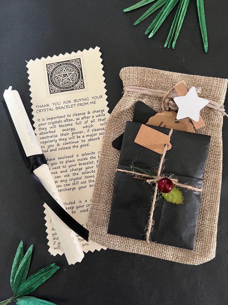 May include: A gift set with a burlap bag, wrapped package, and a scroll. The wrapped package is black with twine and a red berry. The scroll is tied with a black ribbon. The text on the card reads "THANK YOU FOR BUYING YOUR CRYSTAL BRACELET FROM ME."