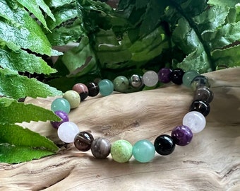 May include: A multi-coloured beaded bracelet with green, purple, brown and white beads. The bracelet is made of natural stones and is photographed on a wooden surface.