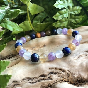 May include: A beaded bracelet featuring a variety of colorful gemstones. The bracelet includes round beads in shades of purple, blue, yellow, white, and brown. The bracelet is displayed on a piece of wood with green foliage in the background.