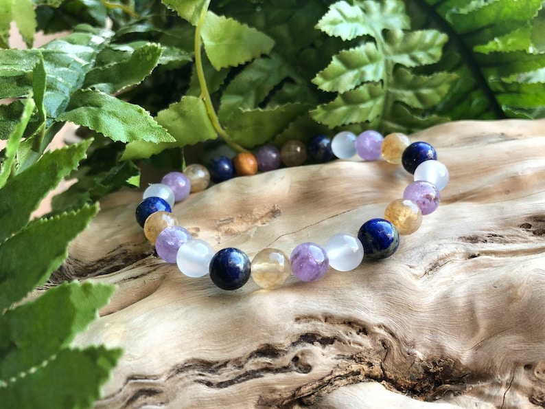 May include: A beaded bracelet featuring a variety of colorful gemstones. The bracelet includes round beads in shades of purple, blue, yellow, white, and brown. The bracelet is displayed on a piece of wood with green foliage in the background.