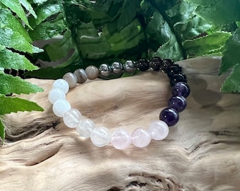 May include: A beaded bracelet with a gradient of colours, starting with white, then pink, then grey, then black, and finally purple. The bracelet is made with natural stones.
