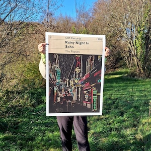 May include: A black and white poster with a neon-coloured image of a rainy street scene in Soho, London. The poster has the text "Stiff Records" and "Rainy Night In Soho" in white text. The text "The Pogues" is in white text below the title. The year "1986" is in white text in the upper right corner.