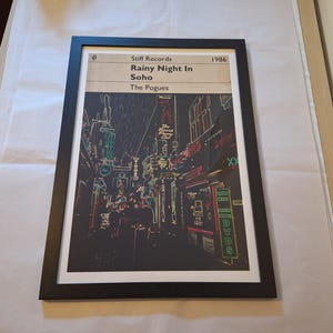 May include: A black and white framed print of a street scene in Soho, London. The print is titled "Rainy Night in Soho" and is by The Pogues. The print is from Stiff Records and is dated 1986.