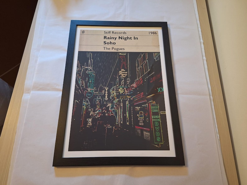 May include: A black and white framed print of a street scene in Soho, London. The print is titled "Rainy Night in Soho" and is by The Pogues. The print is from Stiff Records and is dated 1986.