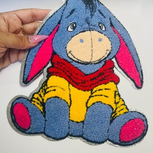 May include: A plush patch of Eeyore, the donkey character from Winnie the Pooh. The patch features a blue body, pink ears and feet, a red scarf, and a yellow shirt. The patch is outlined in silver glitter.