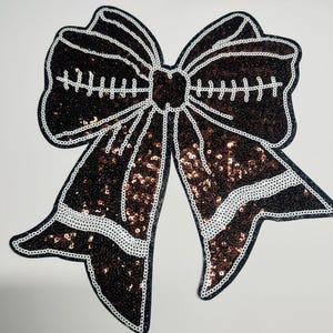 May include: A large, decorative bow applique with a sequined design. The bow features a dark brown color with white accents and a black outline. The sequins add a shimmering effect to the bow's surface, creating a glamorous look.