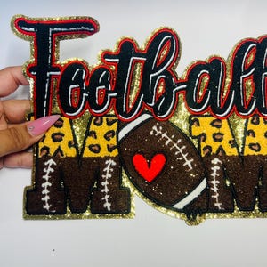 May include: A glittery gold, black, red, and brown "Football Mom" sign. The word "Football" is in black script with red trim. The letters "M" and "M" are brown with white stitching, and the letter "A" has a leopard print design. A football with a red heart is in the center.