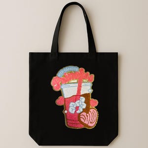 May include: Black tote bag featuring a colorful embroidered design of an iced cafecito. The design includes a coffee cup with ice cubes, a pastry, and the text "ICED CAFECITO" in a blue banner. The bag has black handles.