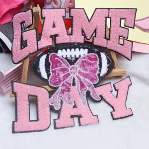 May include: Pink sequined patches spelling "GAME DAY" with a football-shaped patch featuring a sequined bow. The patches have a fuzzy texture and black outlines. The bow and football are embellished with sequins.