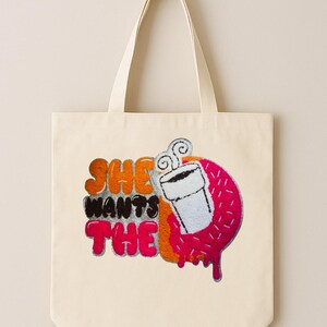 May include: Cream-colored tote bag with the phrase "SHE WANTS THE" in orange, black, and pink lettering. A pink donut with a coffee cup graphic is on the right side. The bag has a simple design with long handles.