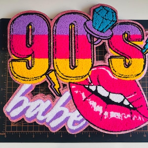 May include: A colorful 90s-themed patch with the word "90s" in large, striped letters. The patch features a diamond-topped hat, a lightning bolt, and a pair of bright pink lips. The word "babe" is also included.