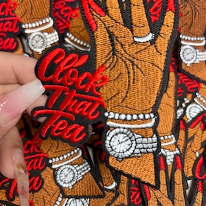 May include: Embroidered patch featuring a brown hand with red fingernails, a silver ring, and a watch. The patch has the words "Clock That Tea" in red script on a black background. The patches are stacked.
