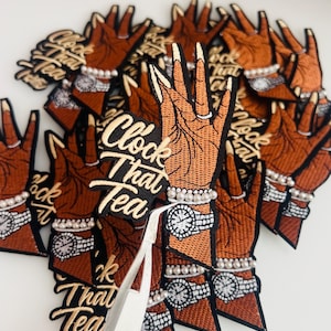 May include: Embroidered patches featuring a brown hand with long, manicured nails, a pearl bracelet, and a watch. The patches have the text "Clock That Tea" in a cursive font. The patches are on a white surface.
