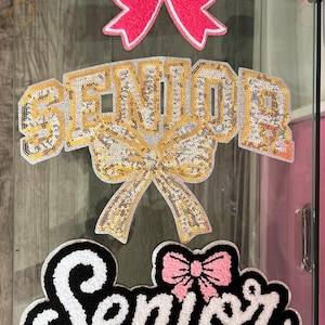 May include: Three decorative signs with the word "SENIOR" in different styles. The top sign is pink with a white outline and a pink bow. The middle sign is gold and silver with a gold bow. The bottom sign is black and white with "2026" in pink.
