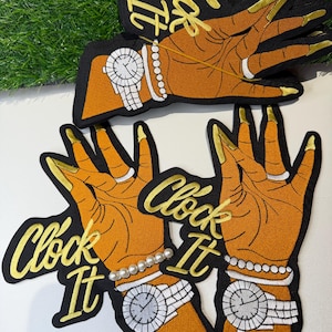 May include: Embroidered patches featuring a hand design with gold fingernails, a wristwatch, and a pearl bracelet. The patches have the words "Clock It" in gold script on a black background. The hand is orange.