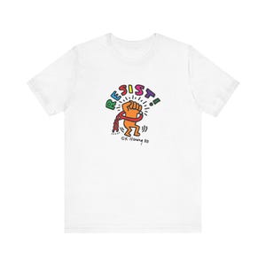 Op de afbeelding: Wit T-shirt met het woord "RESIST" in kleurrijke letters boven een afbeelding van een vuist die kettingen breekt. Het ontwerp bevat de handtekening van de kunstenaar, "©K. Haring 88".