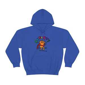 Puede incluir: Una sudadera con capucha azul real con un gráfico colorido. El diseño presenta un puño en alto y la palabra "RESIST!" en colores arcoíris. El gráfico incluye una figura encadenada y la firma del artista, "©K.Haring 88."