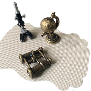 Può includere: Un set in miniatura composto da un microscopio nero e argento, un mappamondo color ottone su base circolare e un binocolo color ottone. Questi tre piccoli oggetti sono disposti insieme su un cartoncino smerlato dalla superficie testurizzata.
