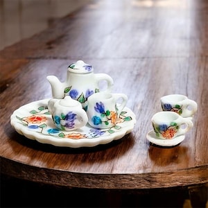 Può includere: Un servizio da tè in miniatura in porcellana bianca con disegni floreali. Il set comprende una teiera, una lattiera, una zuccheriera, due tazze da tè e un vassoio. I fiori sono dipinti in blu, arancione e viola. Il set è esposto su un tavolo di legno scuro.