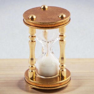 Può includere: Una clessidra con parte superiore e base in legno e dettagli in metallo color oro. Il centro in vetro trasparente contiene sabbia bianca. La clessidra è un oggetto decorativo, probabilmente usato per la misurazione del tempo.