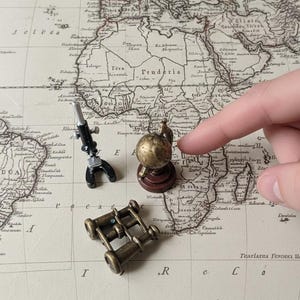 May include: A miniature black and silver microscope, a small brass-colored globe on a dark wooden base, and a pair of antique-style brass binoculars arranged on a vintage-style map featuring geographical outlines and printed text.