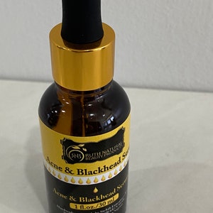 May include: A brown glass bottle with a gold cap and black dropper. The bottle is labeled "Acne & Blackhead Serum" with the brand name "Ruth Natural Beauty Products". The bottle contains 1 fl. oz. (30 ml) of serum.