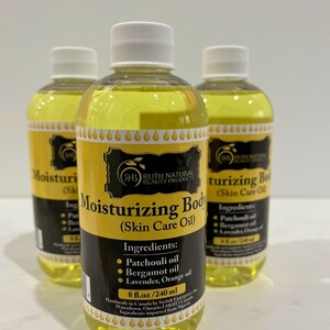 Op de afbeelding: Drie amberkleurige glazen flessen met hydraterende body oil met een geel etiket. Het etiket vermeldt "Moisturizing Body (Skin Care Oil)" en somt de ingrediënten op: patchouli-olie, bergamot-olie, lavendel, sinaasappelolie. De flessen zijn 8 fl. oz. / 240 ml.