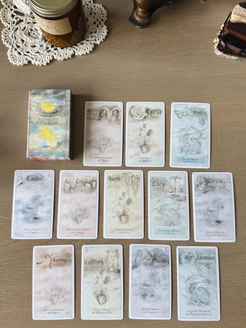 May include: A deck of astrology cards with a box featuring the title "Astrology The Path". The cards are illustrated with zodiac signs, flowers, and symbols. The cards are arranged on a light brown surface.