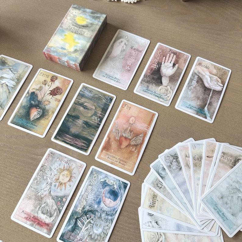 May include: A deck of astrology-themed tarot cards with watercolor illustrations. The cards are spread out on a wooden surface, with the box in the upper left corner. The box reads "Astrology". The cards feature images of hands, hearts, and celestial bodies.