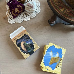 May include: A deck of "Angel Therapy Oracle Cards" by Doreen Virtue sits next to a small stack of cards. The box features an illustration of two figures. A purple velvet bag with gold embroidery and a white doily are also visible.