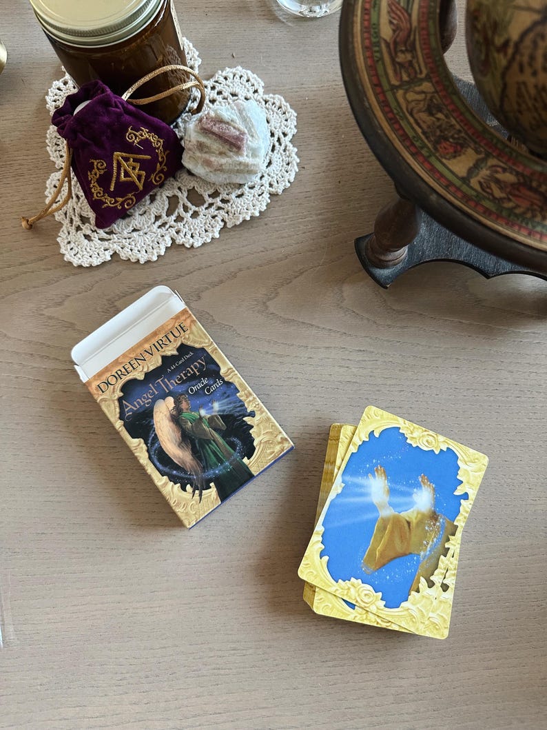 May include: A deck of "Angel Therapy Oracle Cards" by Doreen Virtue sits next to a small stack of cards. The box features an illustration of two figures. A purple velvet bag with gold embroidery and a white doily are also visible.
