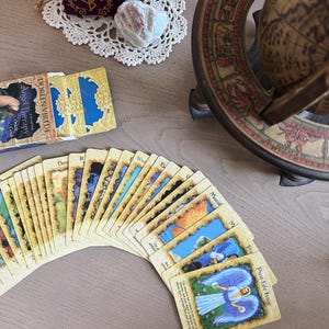 May include: A deck of Doreen Virtue Angel Therapy Oracle Cards fanned out on a light-colored wooden surface. The cards feature colorful illustrations and text. A small purple velvet bag, a white crystal, and a decorative doily are also visible.